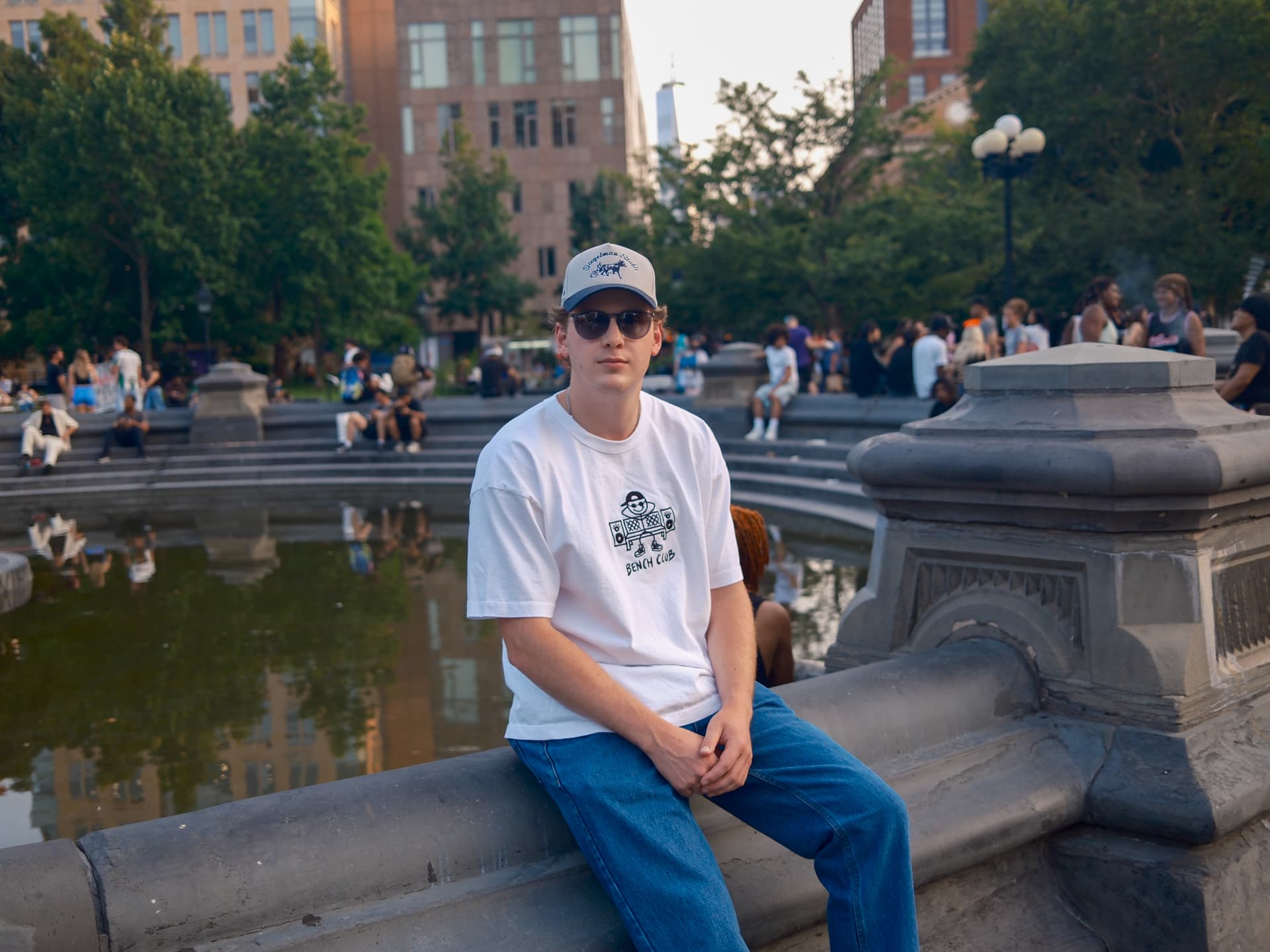 Man in a white Bench Club tee and cap at the Washington Square Park fountain