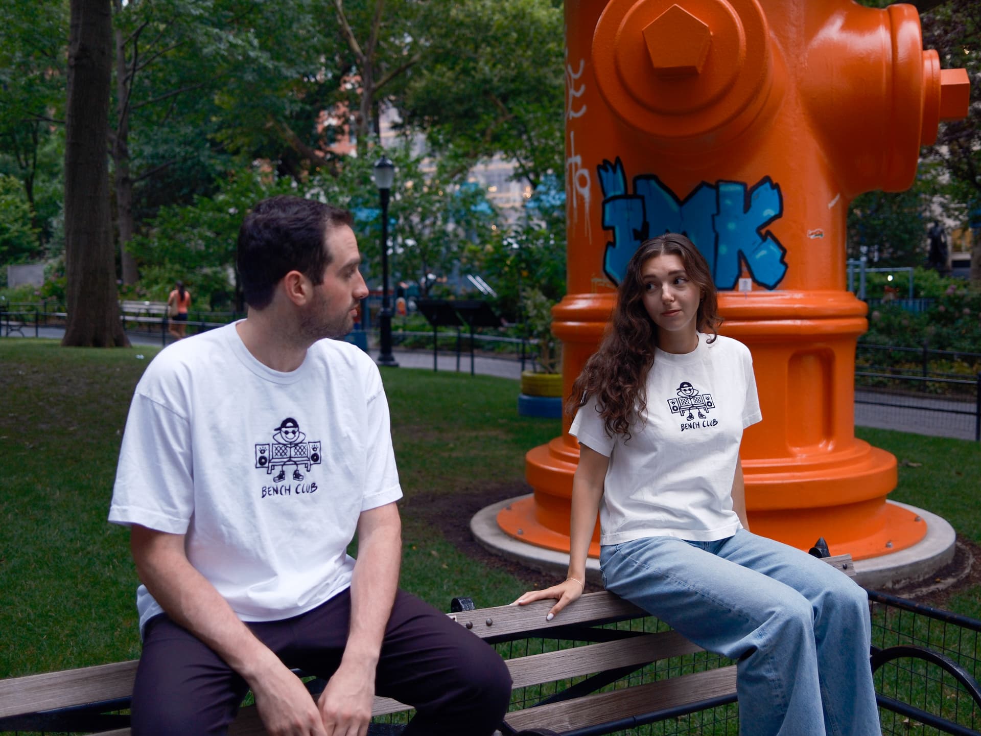 Two people in white Bench Club tees on a park bench by an orange hydrant sculpture