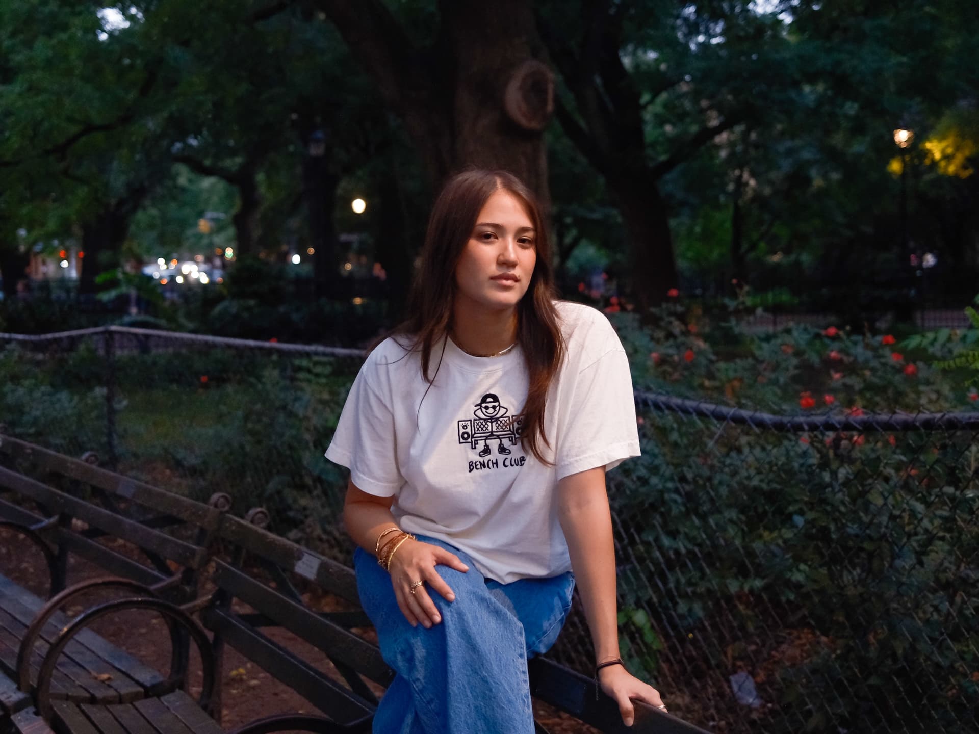 Woman in a white Bench Club tee on a park railing at dusk