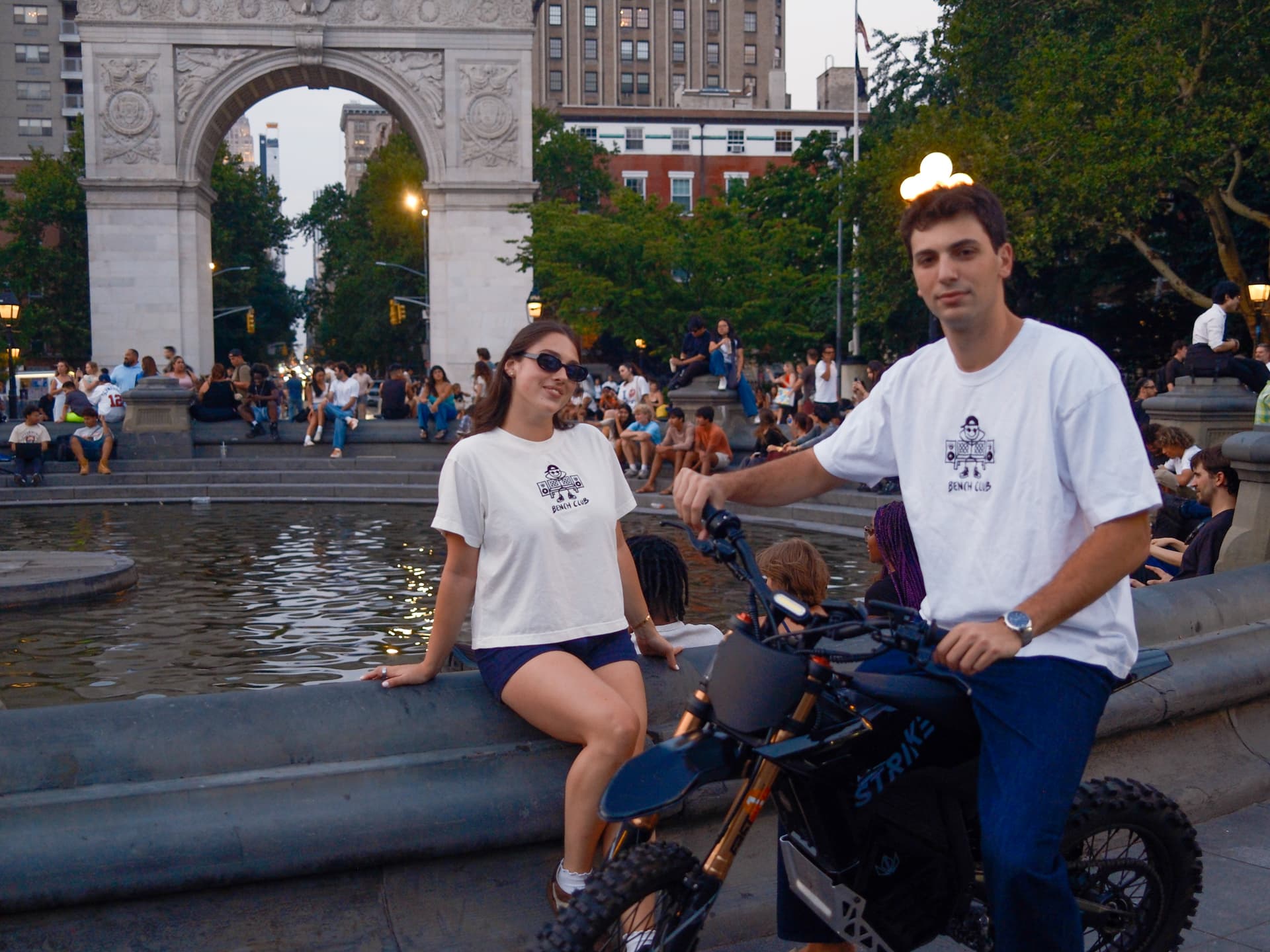 Two people in white Bench Club tees at the Washington Square Park fountain